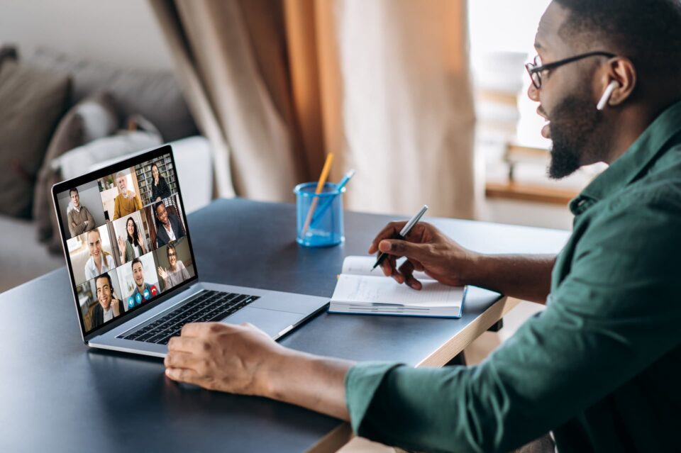A man on a group video call webinar on his laptop while taking notes on some paper
