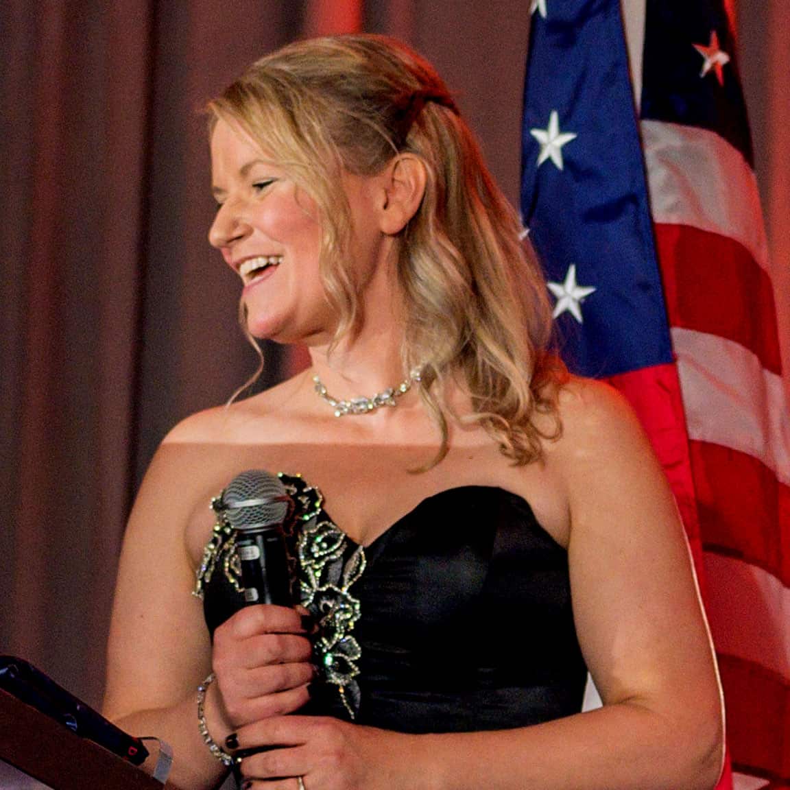 Woman with a microphone smiling at a formal event with American flag in background.