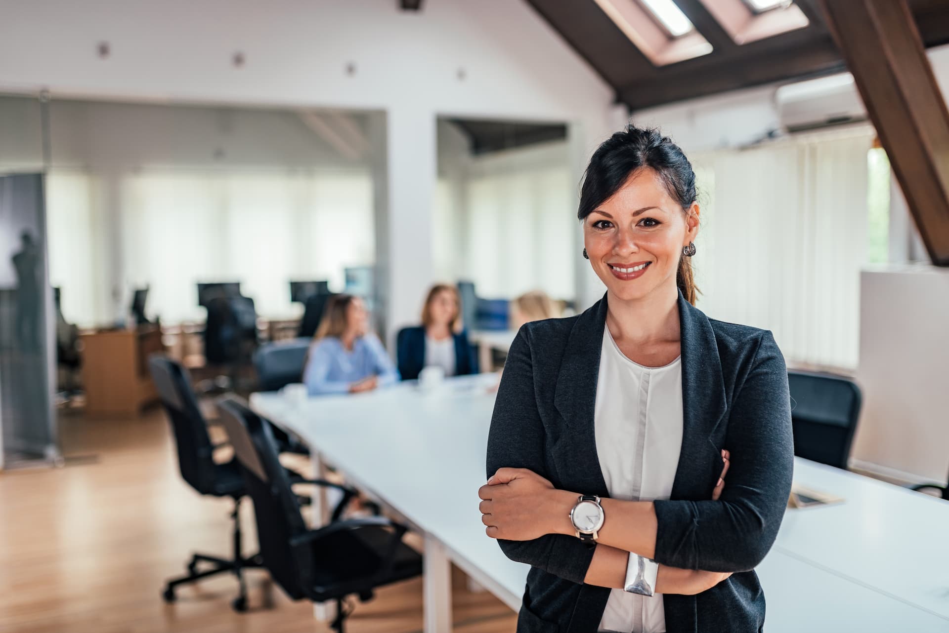 A successful business leader crossing her arms in front of a table that her colleagues are working at