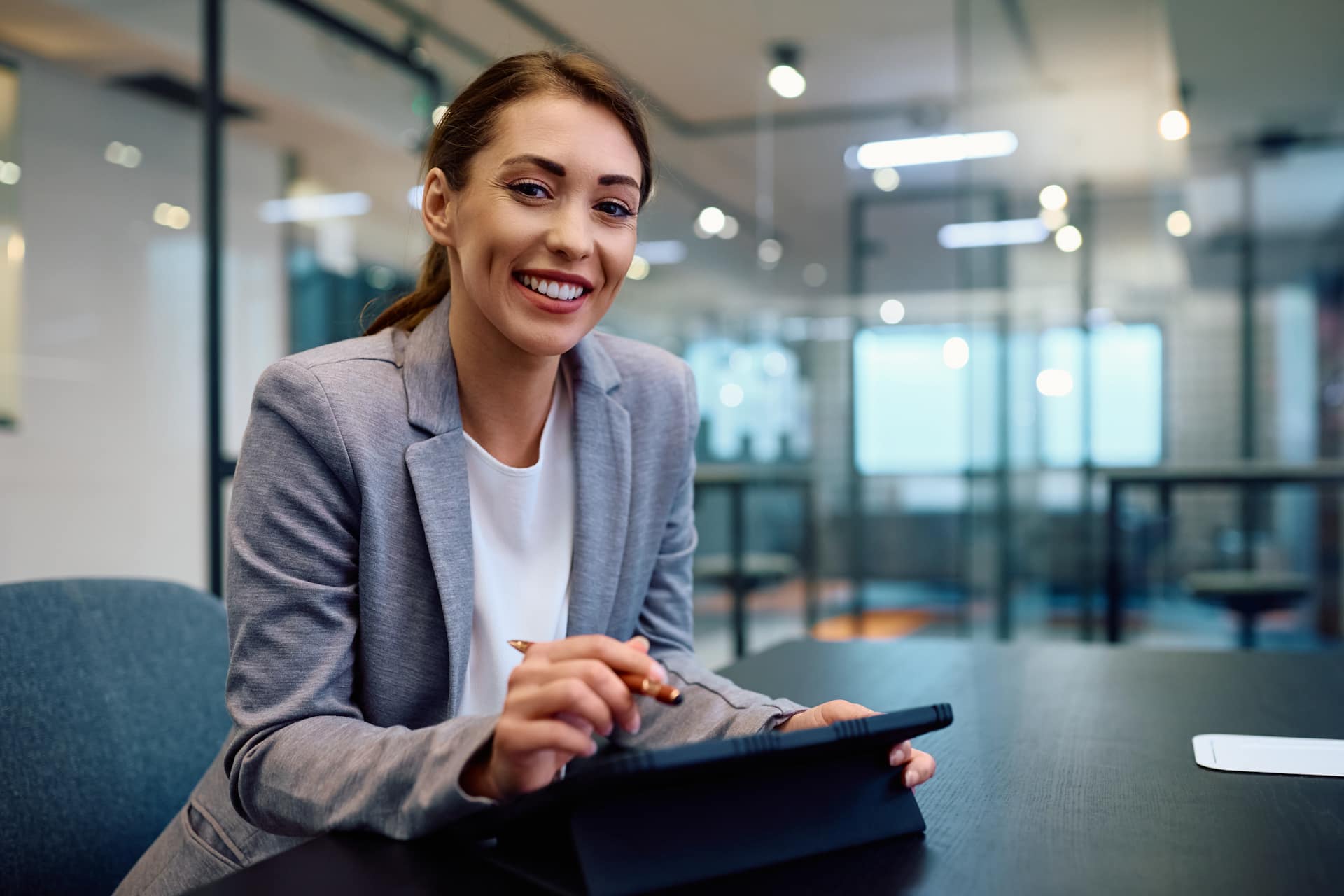 Independent insurance agent in a gray blazer smiling while using a digital tablet in a modern office environment.