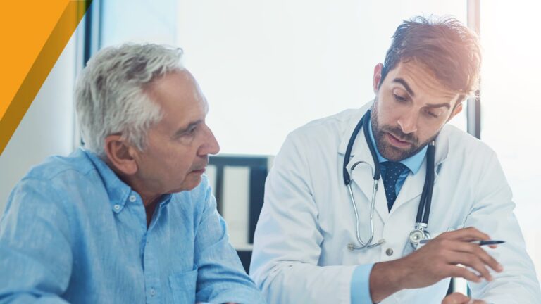 Senior man in light blue shirt consulting with a doctor in white coat and stethoscope who is pointing to documents during a medical appointment in a bright clinic office.