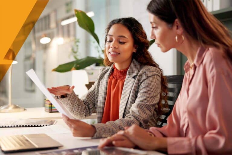 Two businesswomen reviewing a document together at a desk with a laptop in a modern office meeting space.