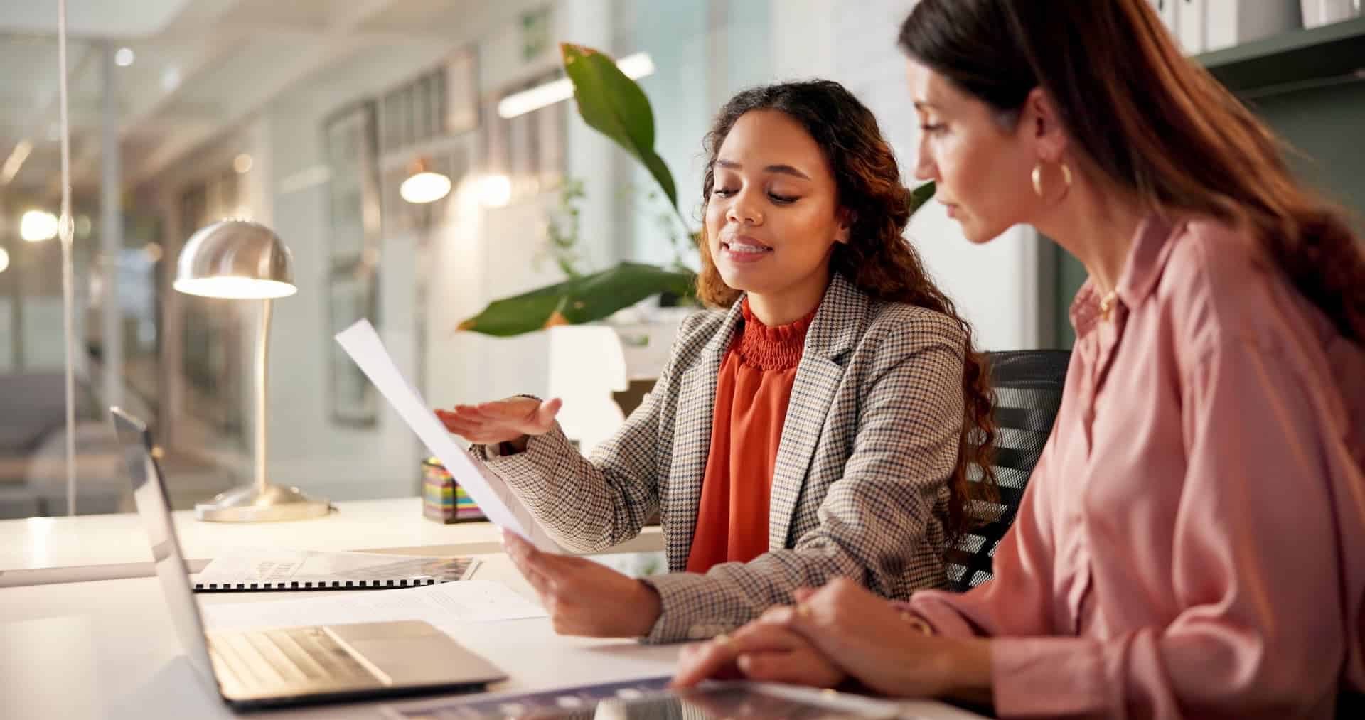 Two businesswomen reviewing a document together at a desk with a laptop in a modern office meeting space.