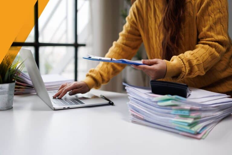 A person managing financial paperwork with a laptop and stacks of documents in an office setting.