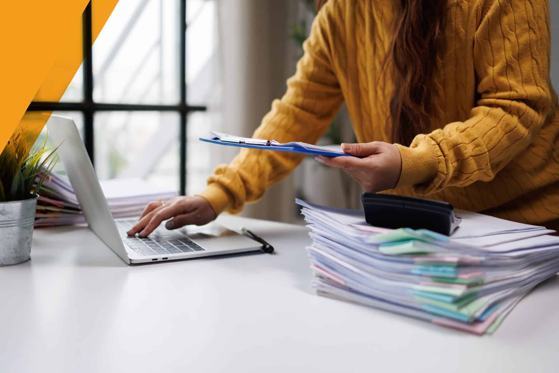 A person managing financial paperwork with a laptop and stacks of documents in an office setting.