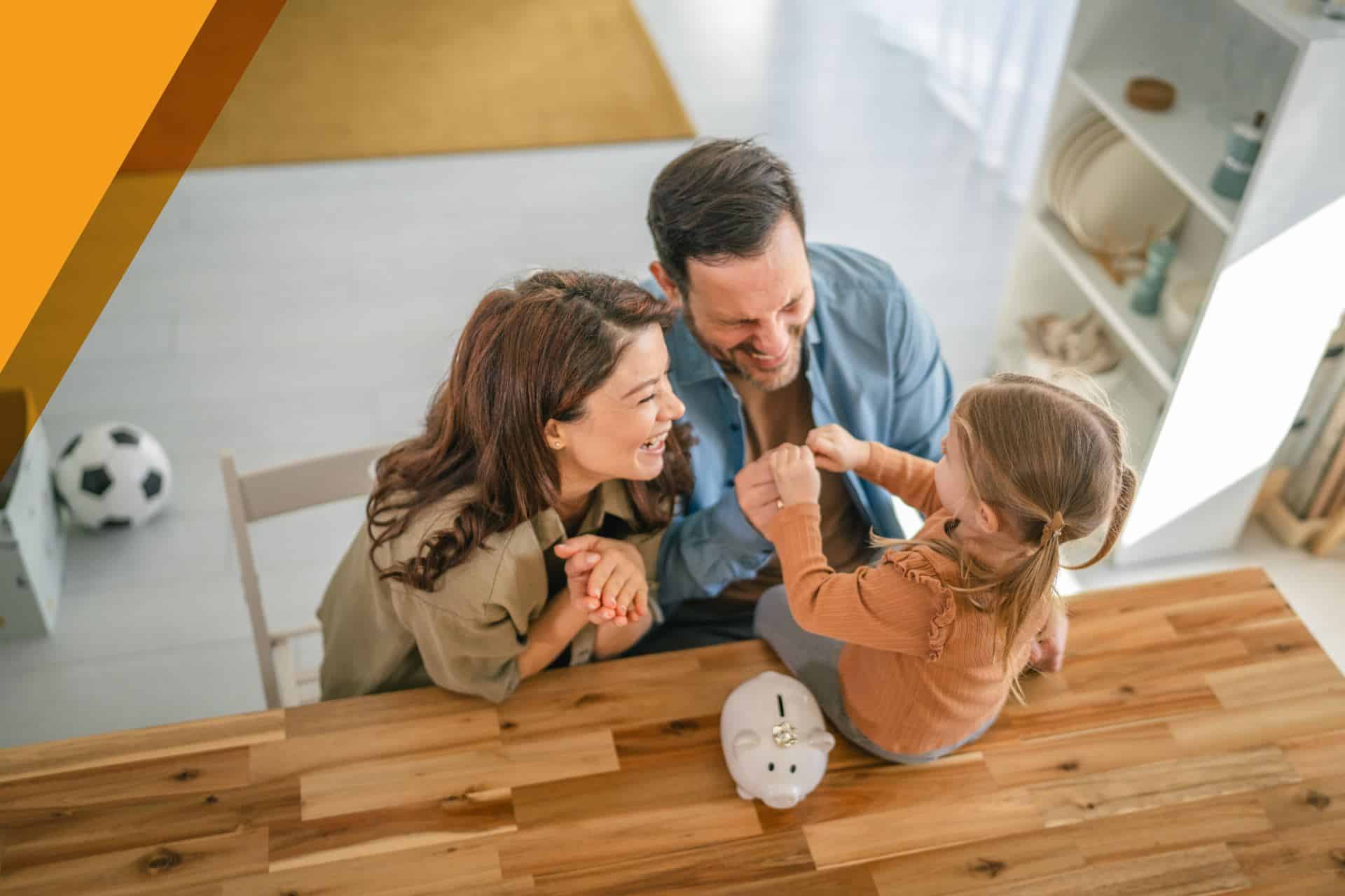 Smiling parents and young daughter holding hands at a wooden table with a piggy bank, saving together at home.